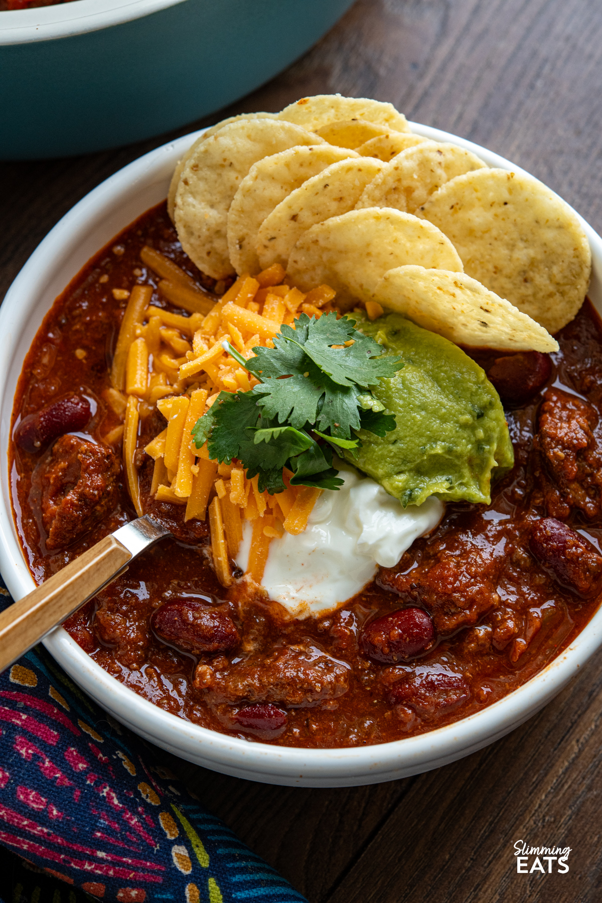 A hearty, flavour-packed slow cooker beef chilli with a rich salsa-infused sauce, tender beans and smoky spices for an easy meal the whole family will love. close up of serving of Slow Cooker Salsa Beef Chilli in bowl with guacamole, soured cream, cheddar, cilantro and tortilla chips