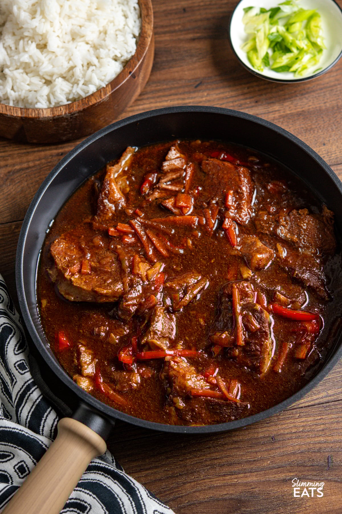 Slow Cooker Honey Tamarind Pork in a skillet with wooden bowl of rice and small bowl of chopped spring onions above.