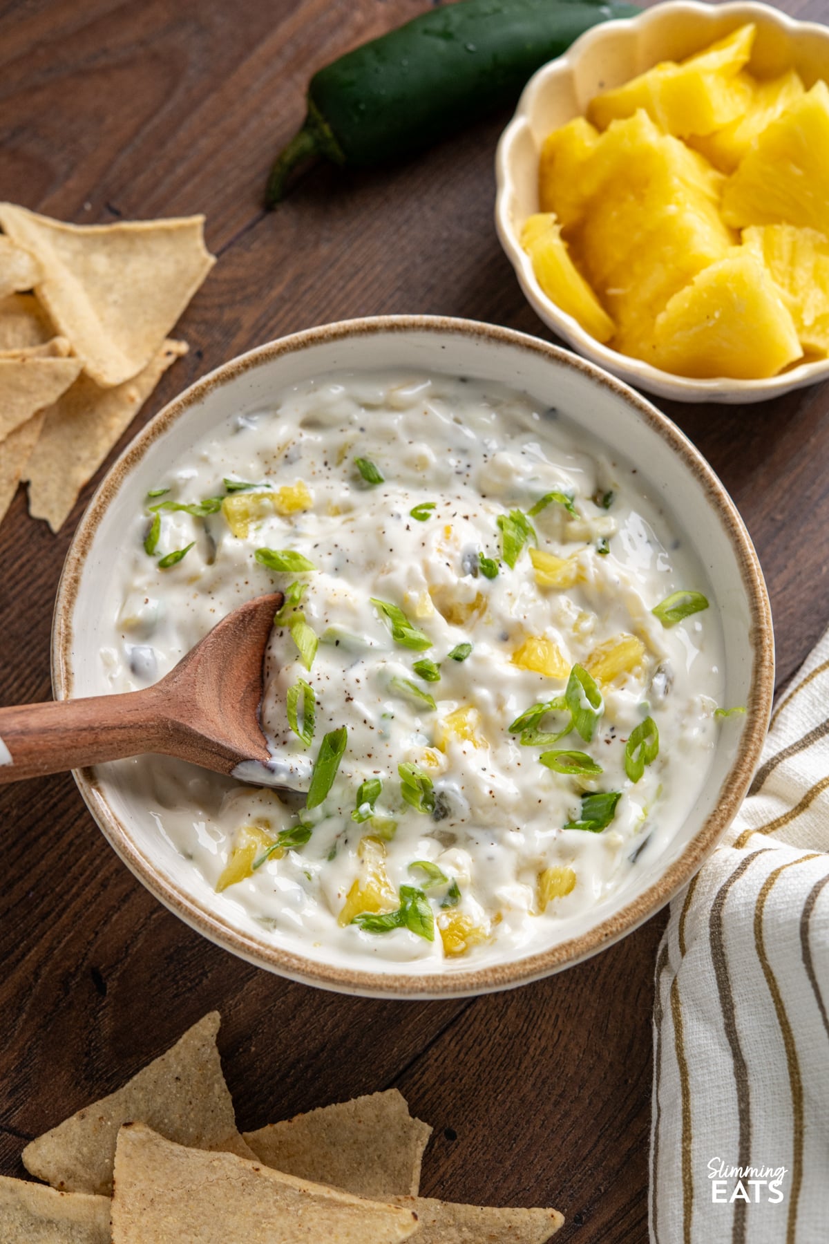 Creamy Jalapeno Pineapple Dip in white bowl with beige rim, wooden spoon placed in dip, surrounded by a bowl with fresh pineapple, tortilla chips and a fresh jalapeno