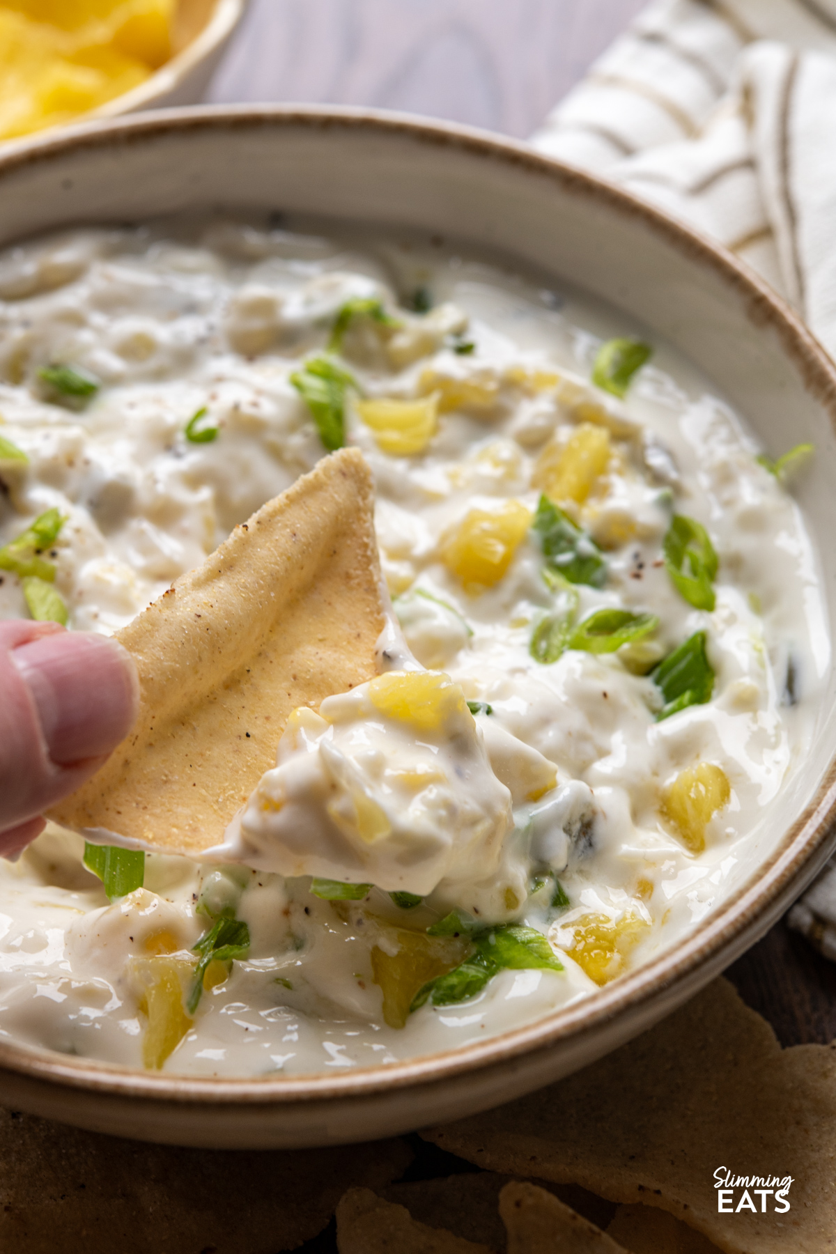 tortilla chip being dipped into creamy jalapeno pineapple dip in a small white bowl with beige rim. 