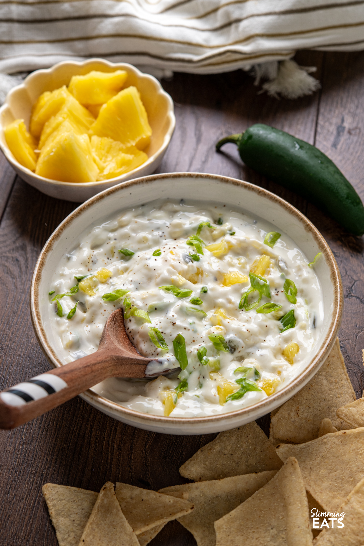 Creamy Jalapeno Pineapple Dip in white bowl with beige rim, wooden spoon placed in dip, surrounded by a bowl with fresh pineapple, tortilla chips and a fresh jalapeno
