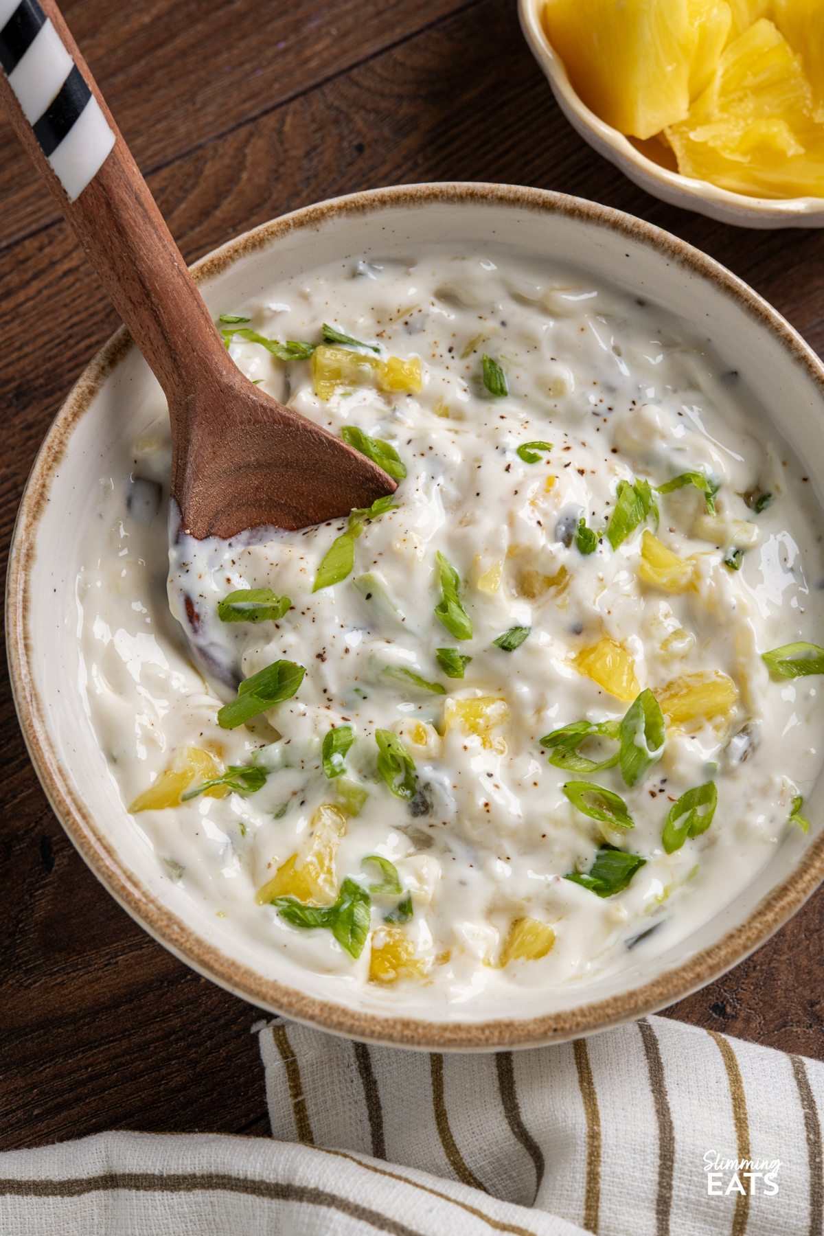 Creamy Jalapeno Pineapple Dip in white bowl with beige rim, wooden spoon placed in dip, surrounded by a bowl with fresh pineapple