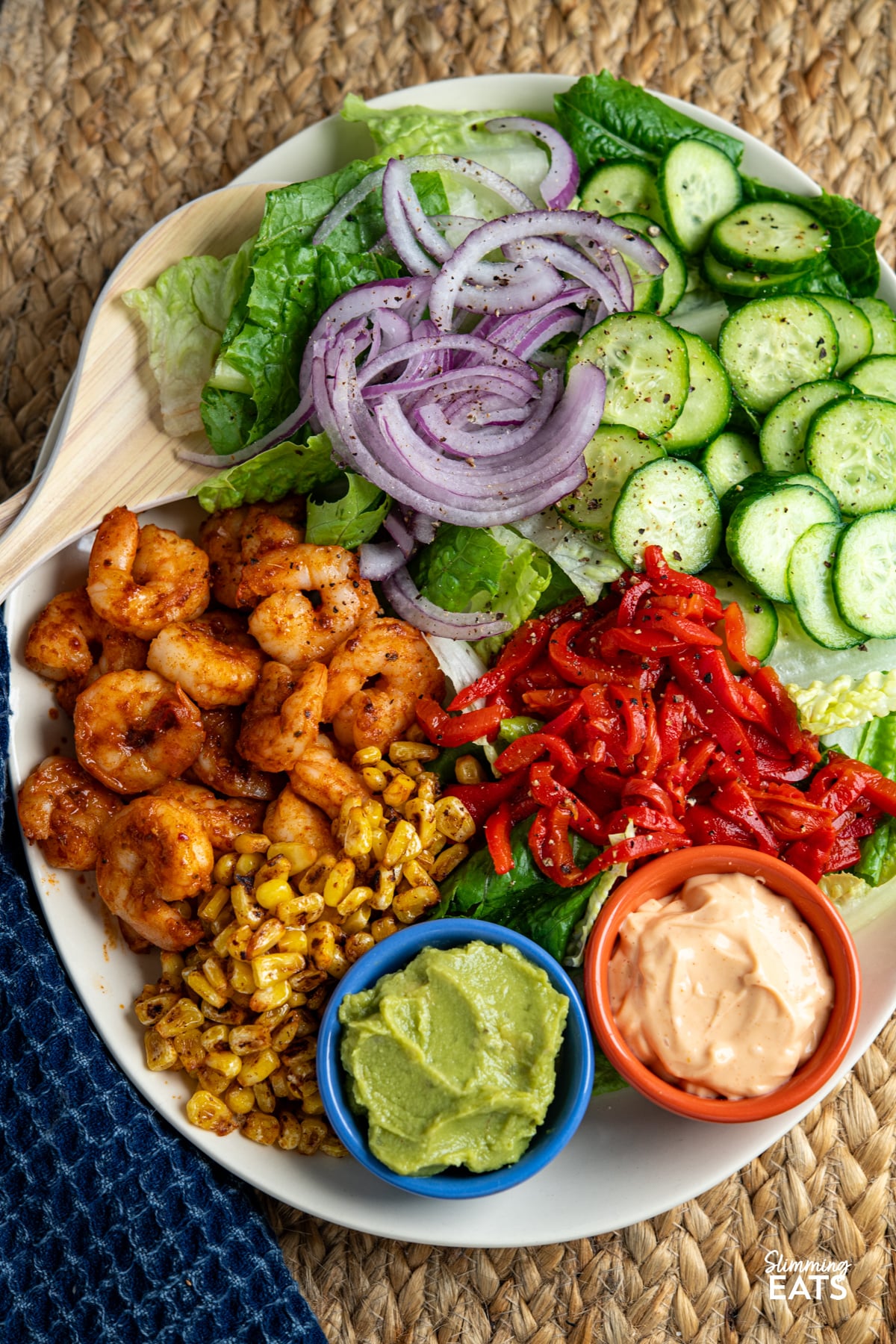Peri Peri King Prawn Salad on a oval plate with salad serving spoons (prawns, lettuce, onion, cucumber, red pepper, charred corn, perinaise and guacamole