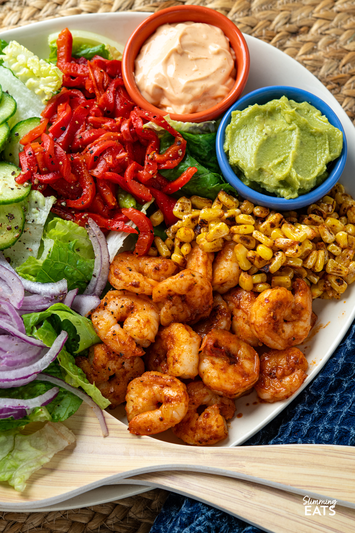 close up of Peri Peri King Prawn Salad on a oval plate with salad serving spoons (prawns, lettuce, onion, cucumber, red pepper, charred corn, perinaise and guacamole