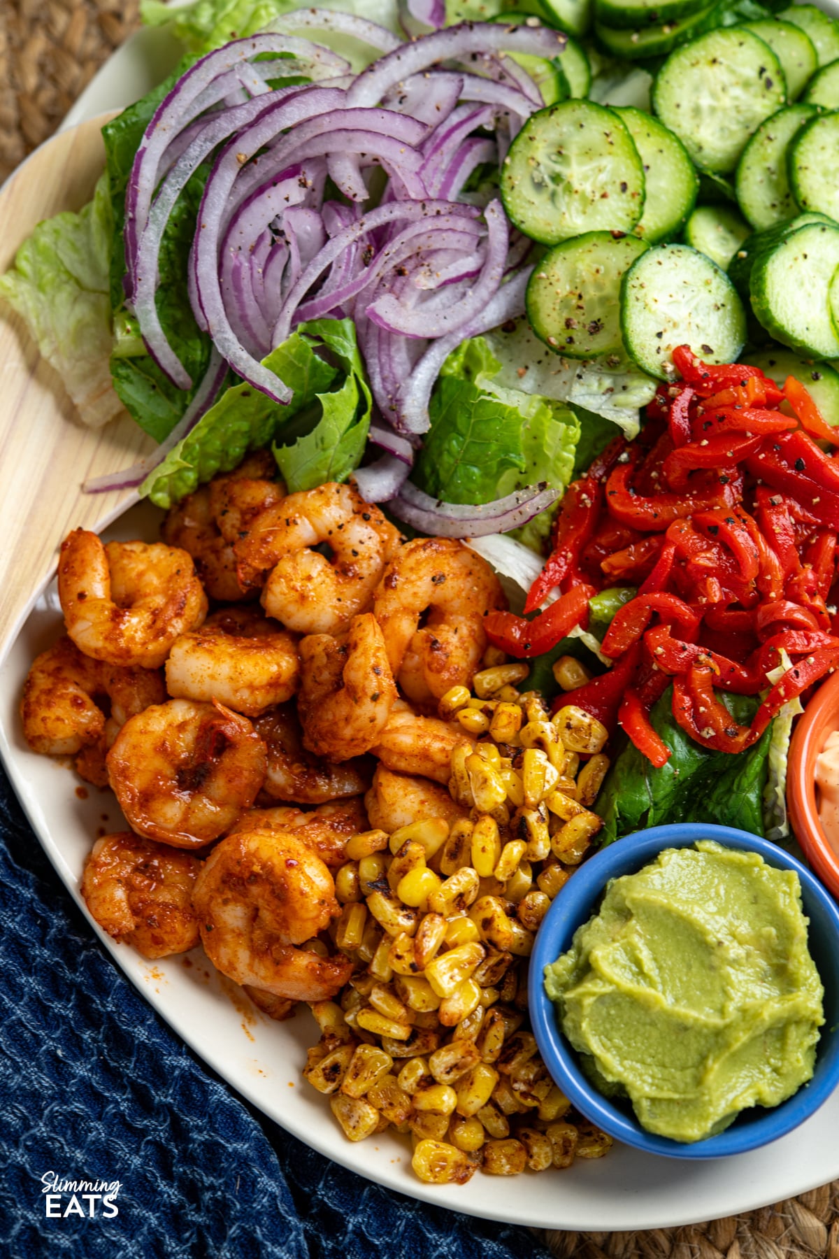 close up of Peri Peri King Prawn Salad on a oval plate with salad serving spoons (prawns, lettuce, onion, cucumber, red pepper, charred corn, perinaise and guacamole