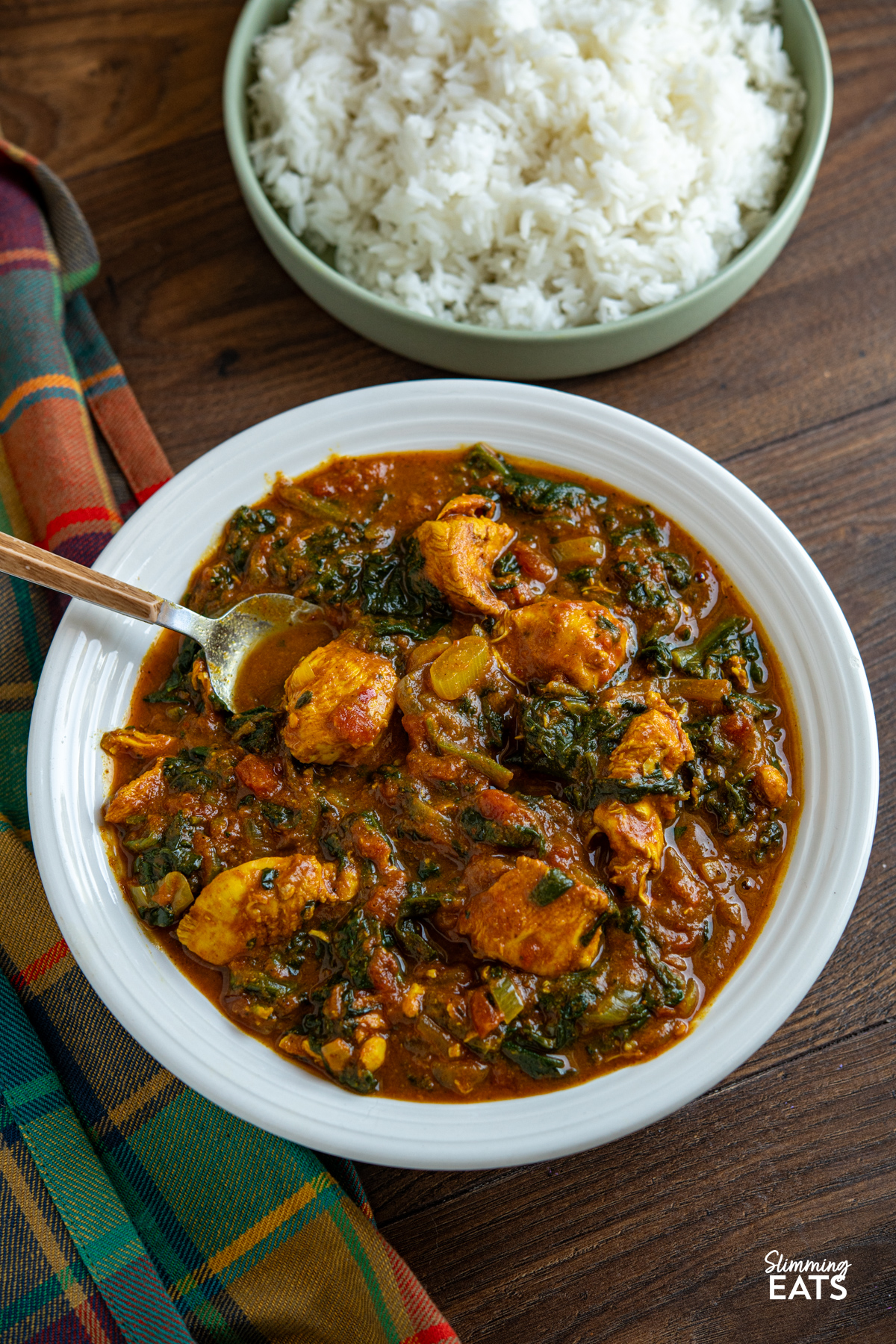 This slow cooker chicken spinach curry is an easy, delicious meal that is perfect for busy days. Tender chicken cooks away in a rich tomato curry sauce, with spinach stirred through at the end for a tasty family-friendly dish that is full of flavour. Slow Cooker Easy Chicken Spinach Curry in a white bowl with a light green bowl of rice