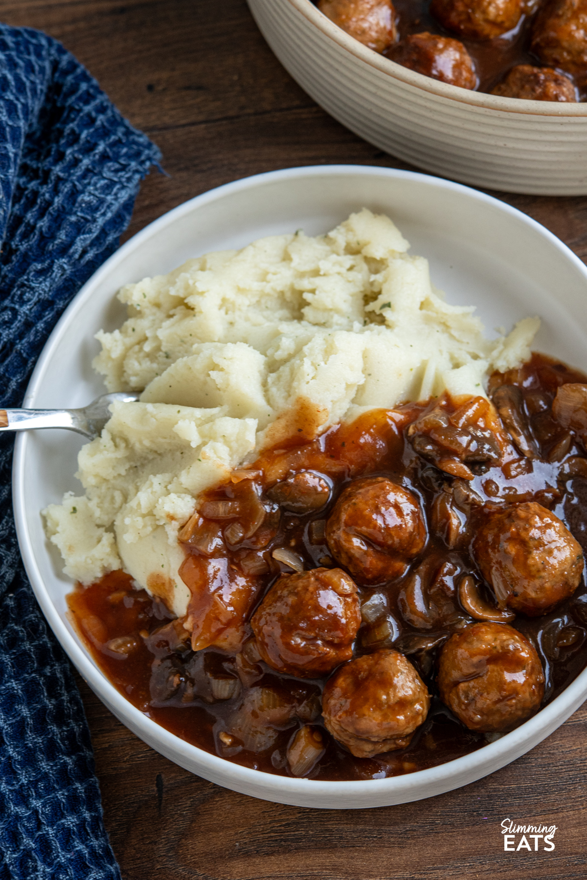 These slow cooker Salisbury meatballs are an easy, hearty dinner made with frozen meatballs simmered in a rich onion and mushroom gravy. close up of serving of Slow Cooker Salisbury Meatballs in a white bowl with mashed potatoes, fork place on left in bowl