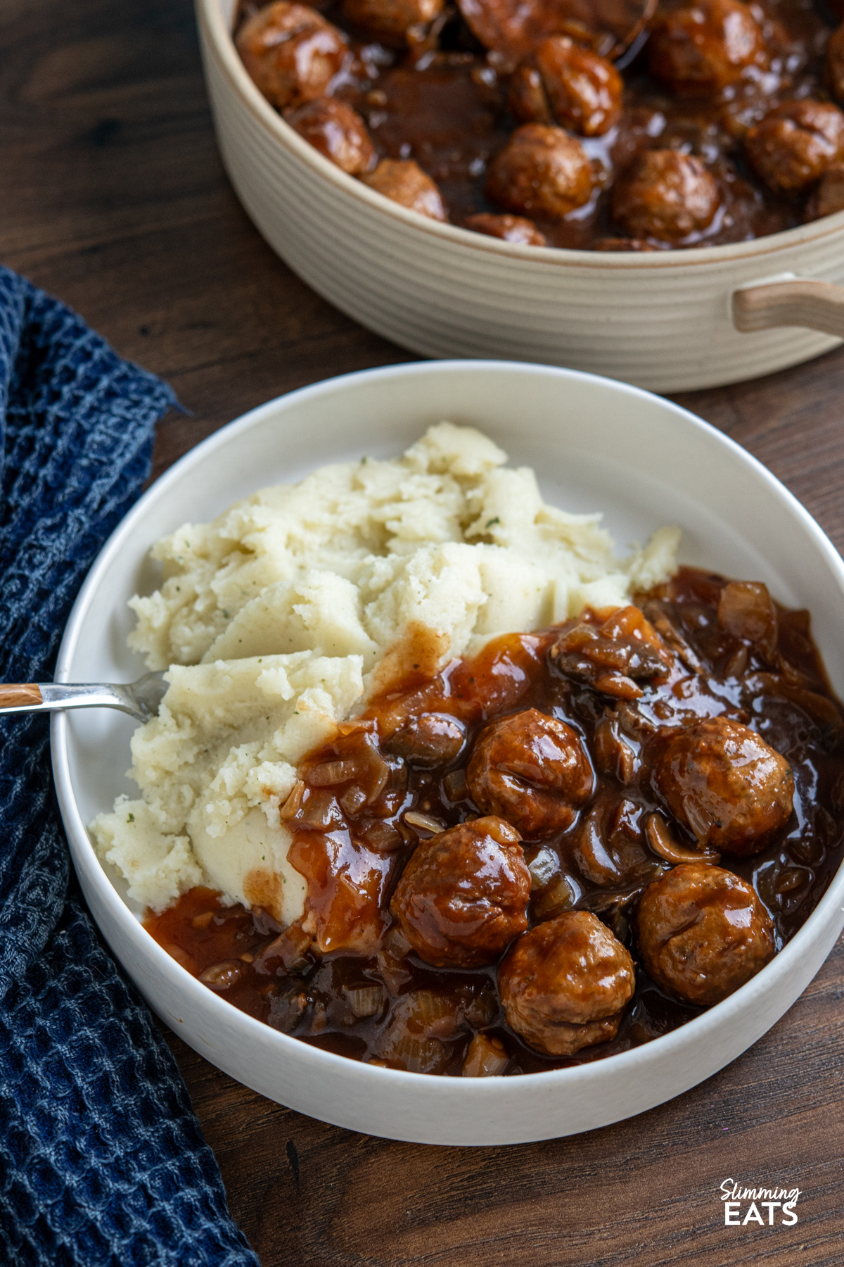 These slow cooker Salisbury meatballs are an easy, hearty dinner made with frozen meatballs simmered in a rich onion and mushroom gravy. serving of Slow Cooker Salisbury Meatballs in a white bowl with mashed potatoes, fork place on left in bowl