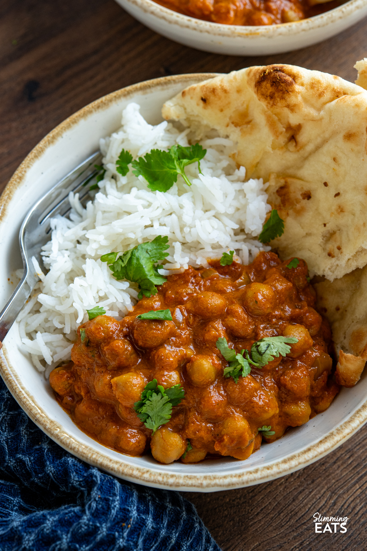 close up of serving of  Slow Cooker Tomato Chickpea Curry in a white bowl with beige rim with white rice and torn naan, garnished with coriander