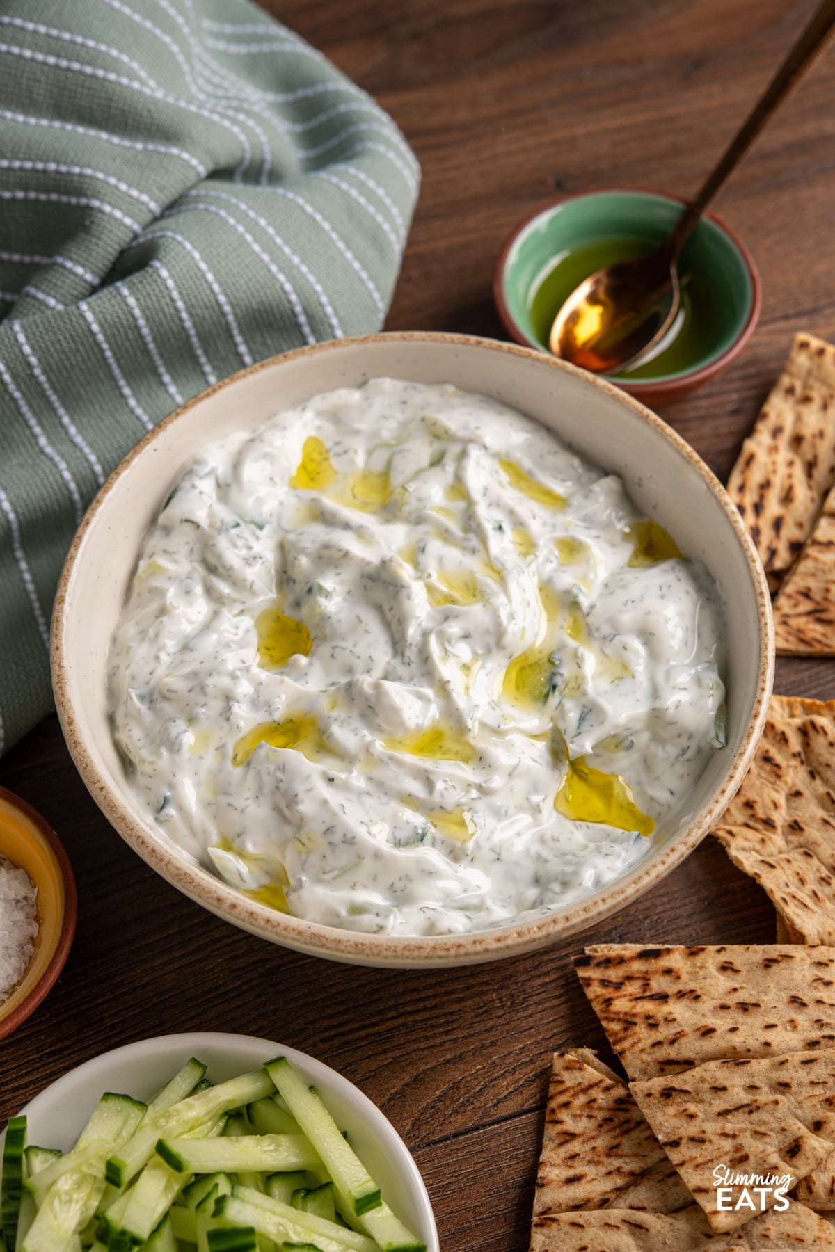 This easy tzatziki dip is cool, creamy and packed with fresh flavour, making it perfect for dipping, drizzling or serving alongside all your favourite Mediterranean-inspired dishes. Easy Tzatziki Dip in a bowl surrounded by pitta and bowls with salt, cucumber and olive oil