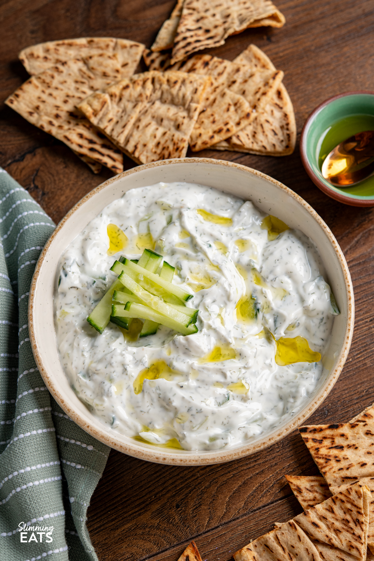 This easy tzatziki dip is cool, creamy and packed with fresh flavour, making it perfect for dipping, drizzling or serving alongside all your favourite Mediterranean-inspired dishes. Easy Tzatziki Dip in a bowl surrounded by pitta and bowls with olive oil