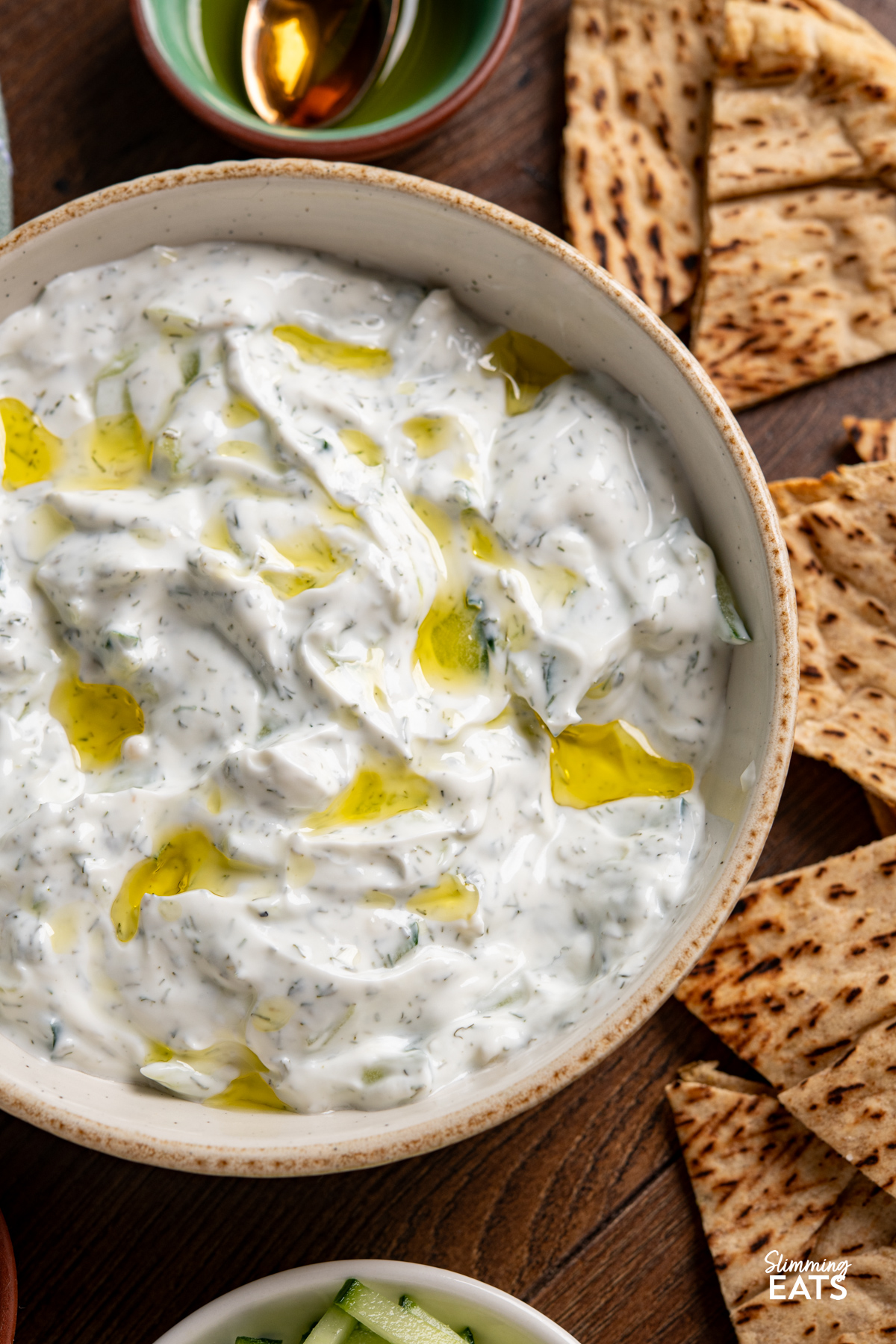 This easy tzatziki dip is cool, creamy and packed with fresh flavour, making it perfect for dipping, drizzling or serving alongside all your favourite Mediterranean-inspired dishes. close up of Easy Tzatziki Dip in a bowl surrounded by pitta and bowls with salt, cucumber and olive oil