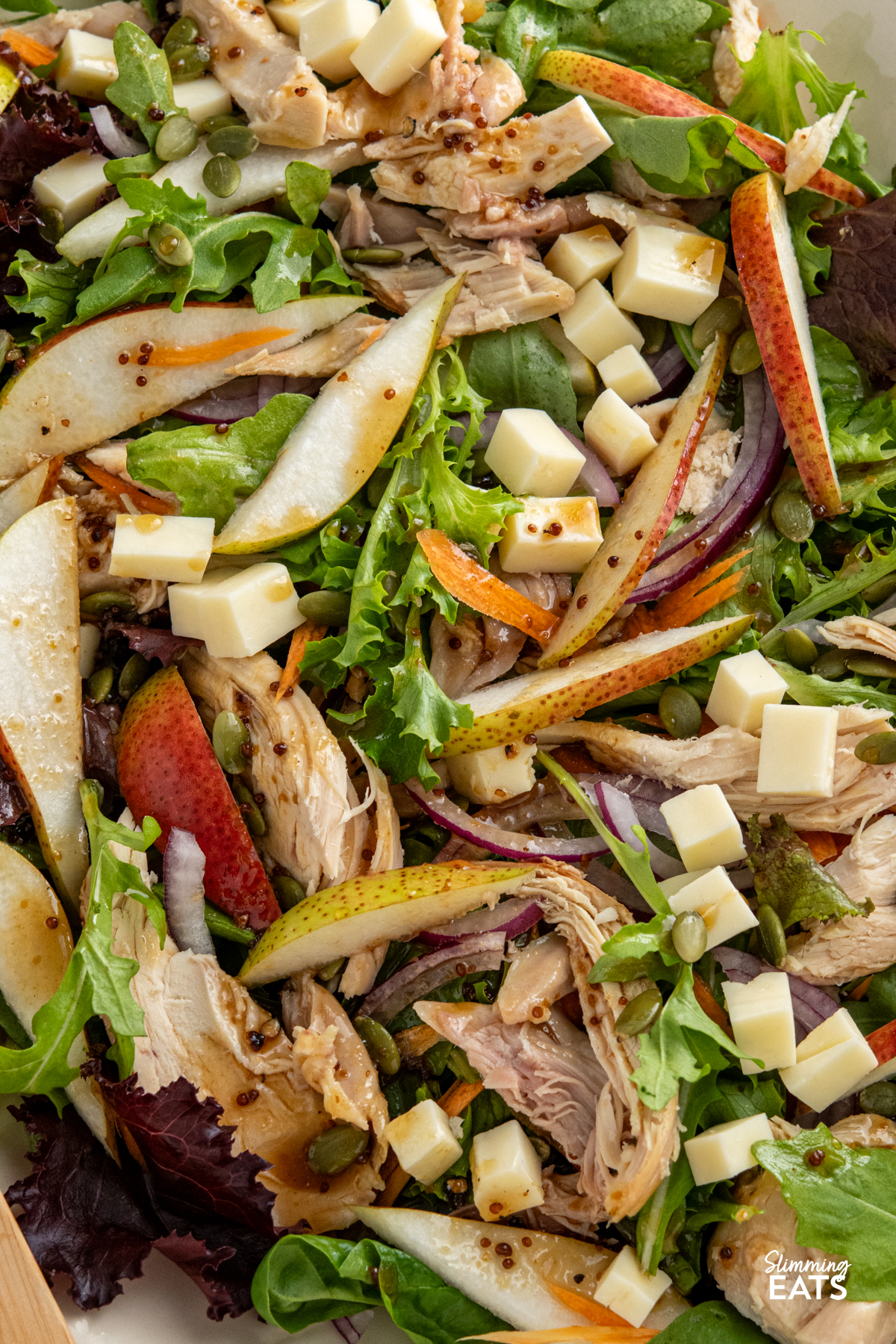 This Chicken Gouda Pear Salad is a delicious mix of sweet juicy pear, gouda, tender chicken, crunchy pumpkin seeds and baby greens, all tossed together with a mustard, lemon and maple dressing. It’s light, colourful and packed with flavour, while still being filling enough for lunch or dinner. close up of Chicken, Gouda and Pear Salad on a oval beige plate with wooden salad spoons drizzled with dressing
