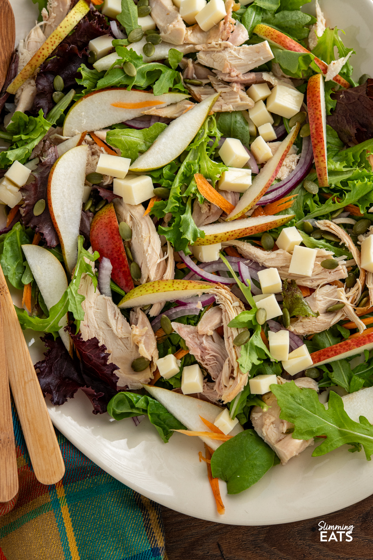 This Chicken Gouda Pear Salad is a delicious mix of sweet juicy pear, gouda, tender chicken, crunchy pumpkin seeds and baby greens, all tossed together with a mustard, lemon and maple dressing. It’s light, colourful and packed with flavour, while still being filling enough for lunch or dinner. close up of Chicken, Gouda and Pear Salad on a oval beige plate with wooden salad spoons