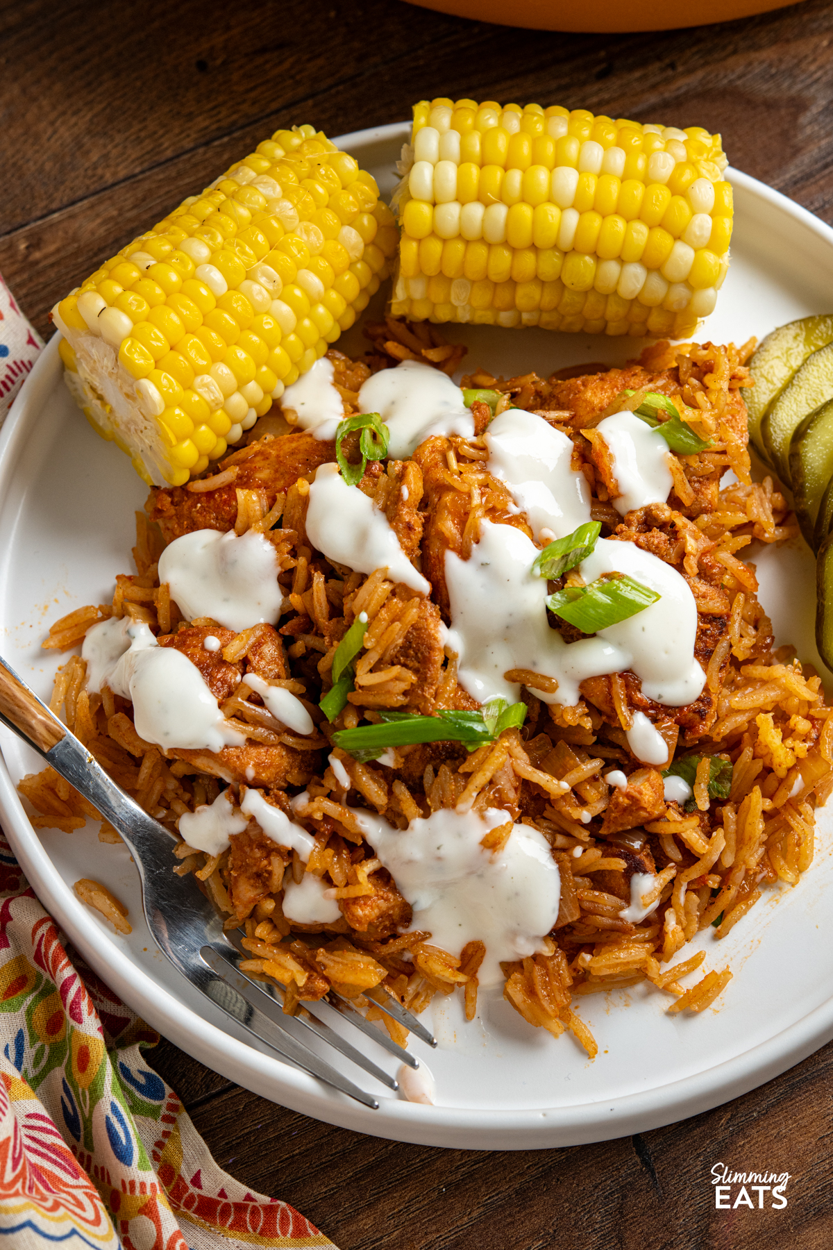 close up of serving of One Pan Nashville Chicken and Rice on white plate drizzled with ranch and served with a side of corn on cob and pickles