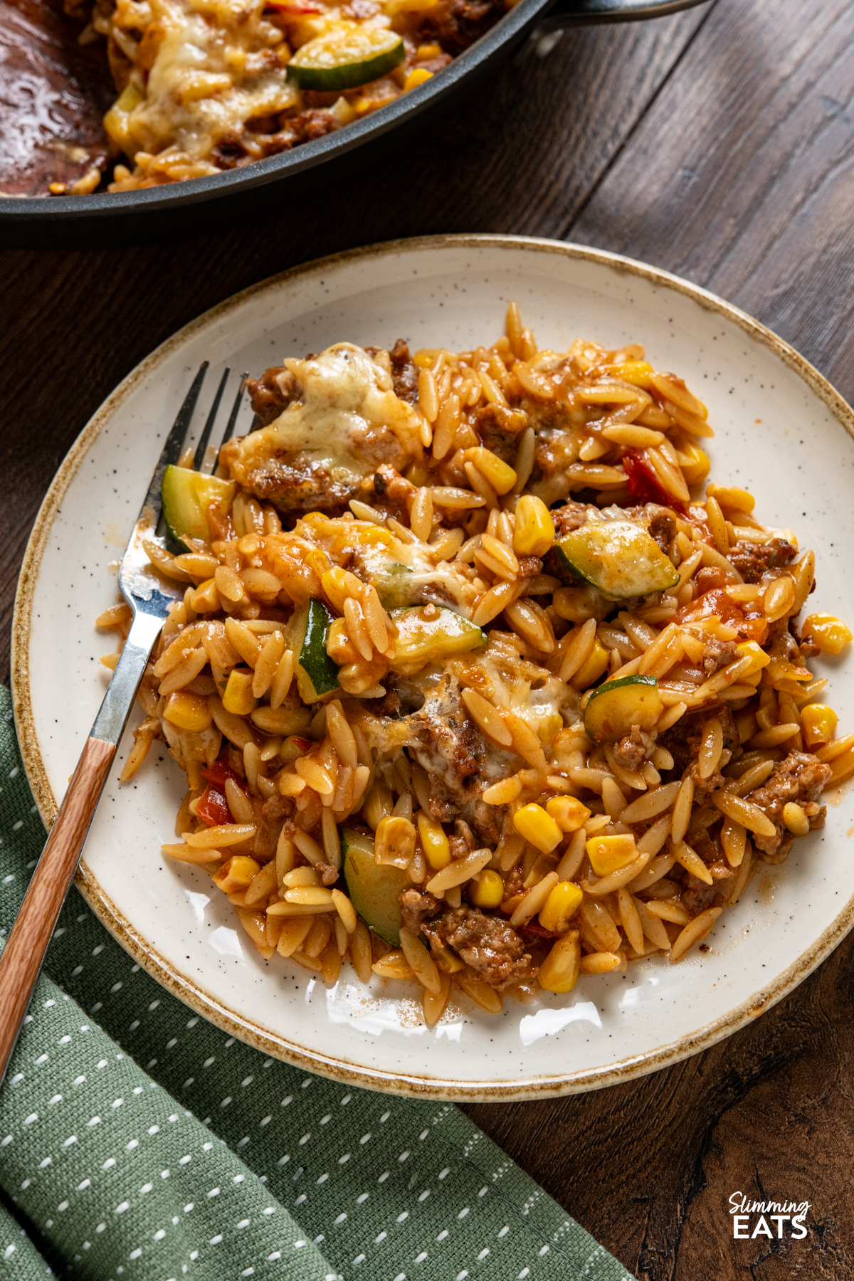 close up of serving of Cheesy Ground Chicken Orzo with Zucchini and Corn one white plate with beige rim, fork with wooden style handle place on plate. 