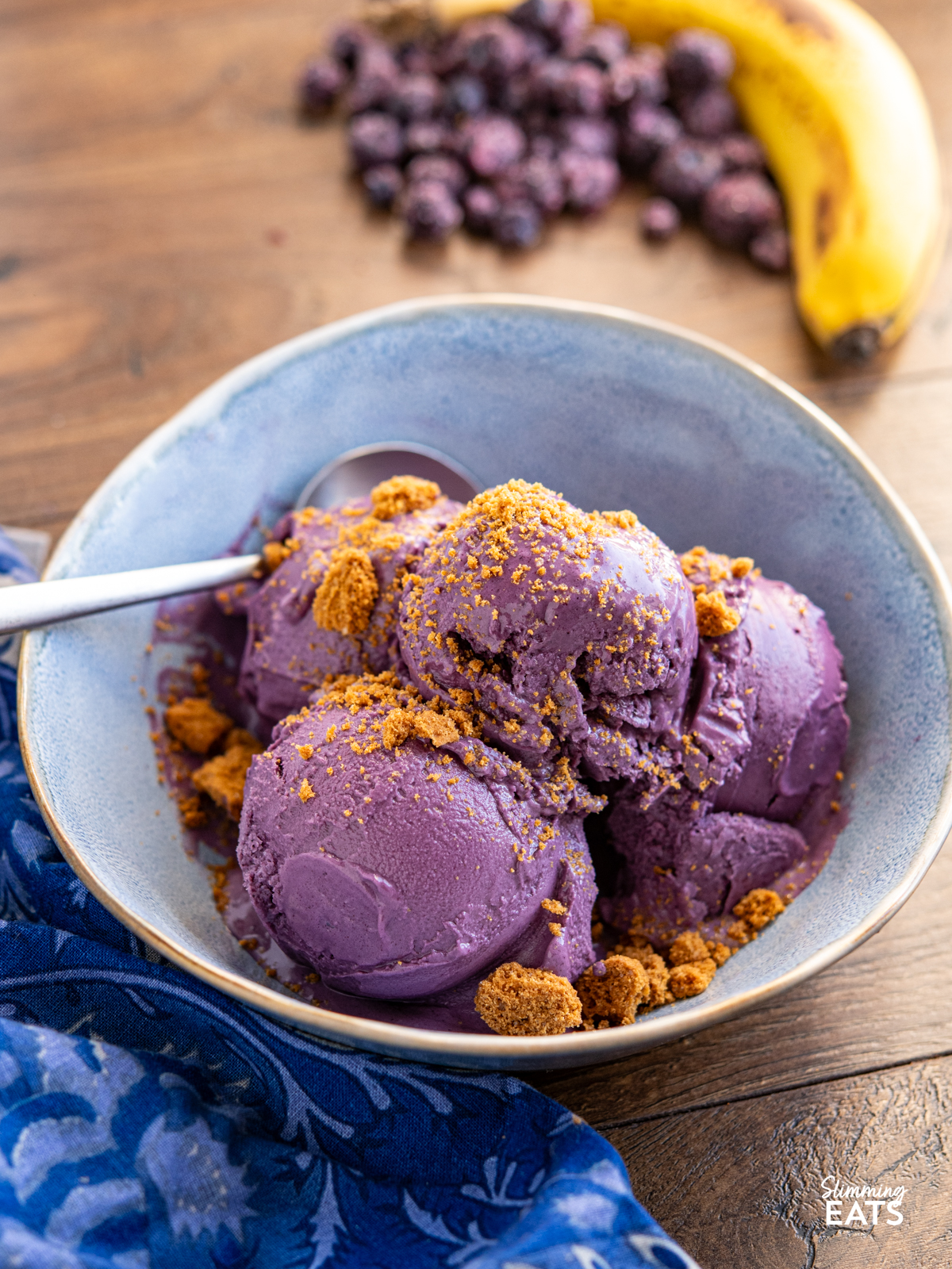 Blueberry Banana Frozen Yoghurt in a blue bowl with spoon topped with crumbled lotus cookie